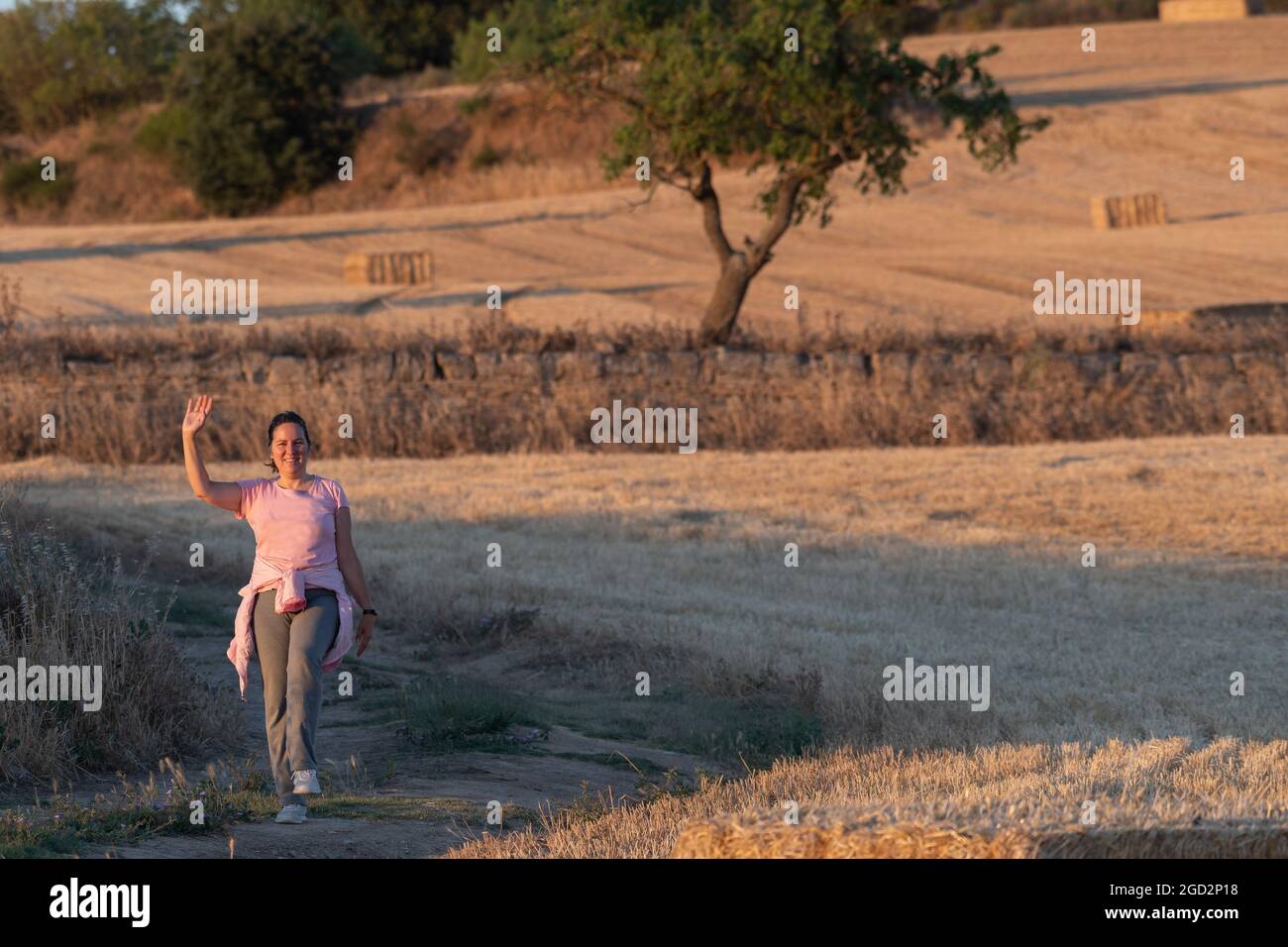 Latina mittleren Alters und eine brünette Frau in rosa und grauer Sportkleidung, die auf dem Land in ländlicher Umgebung spazieren und winken. Selektiver Fokus. Schau Mal Stockfoto