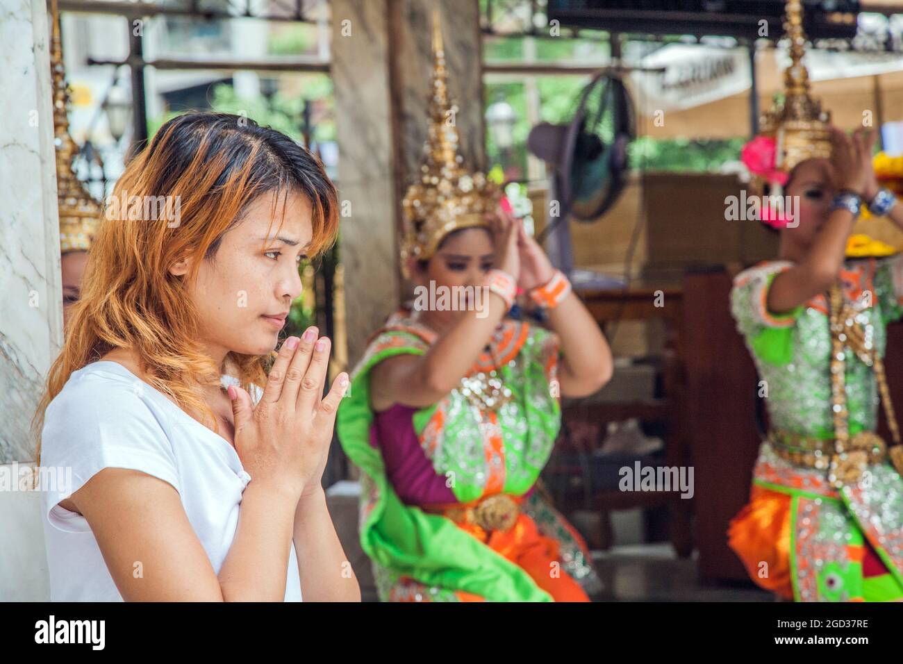 Attraktive Thai-Frau, die im Erawan-Schrein betet, mit prunkvollen Thai-Tänzern im Hintergrund, Bangkok, Thailand Stockfoto