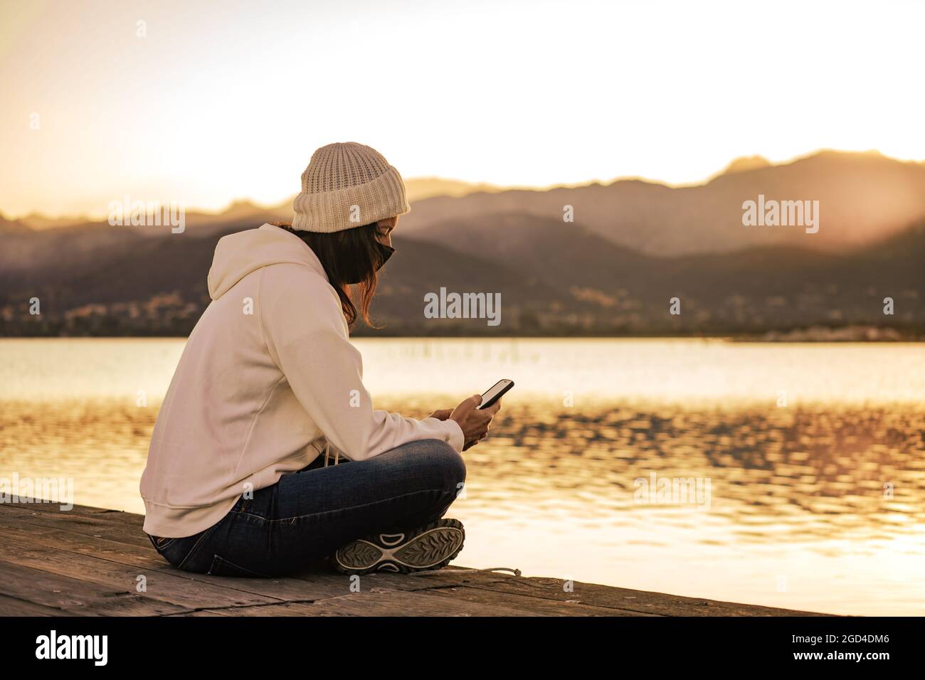 Nicht erkennbare junge Frau in weißem Wollhut sitzt auf einem Pier allein tragen Schutzmaske mit Smartphone in Kontakt mit Freunden und f zu halten Stockfoto
