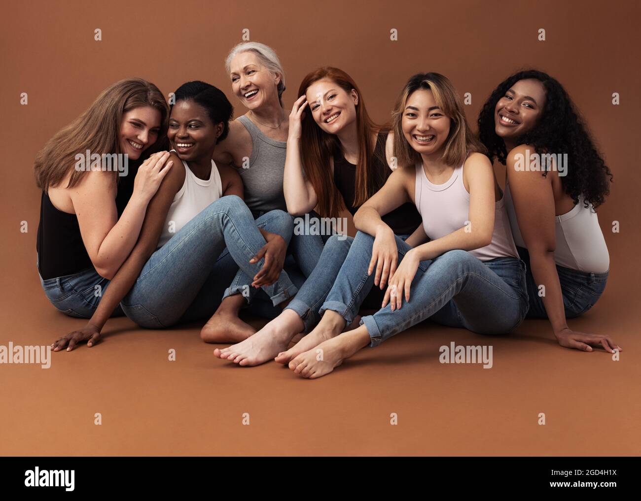 Sechs Frauen unterschiedlichen Alters sitzen zusammen im Studio auf braunem Hintergrund. Multiethnische Gruppe von verschiedenen Frauen, die gute Zeiten haben. Stockfoto