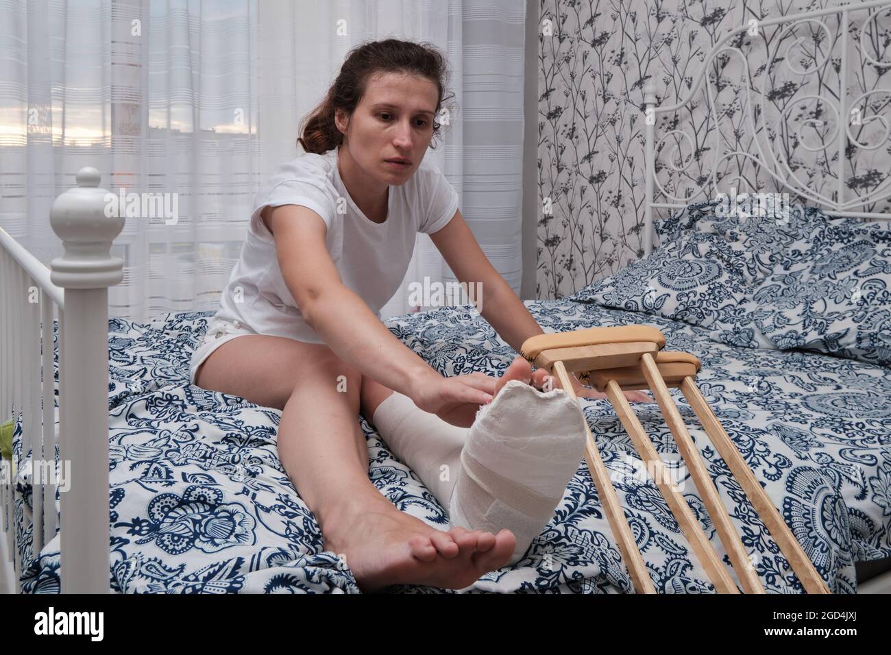 Eine Frau massiert die Zehen eines gebrochenen Beins mit ihren Händen. Draufsicht auf ein gebrochenes Bein in einem Gipsabguss und Krücken. Heimrehabilitation nach einer gebrochenen Stockfoto