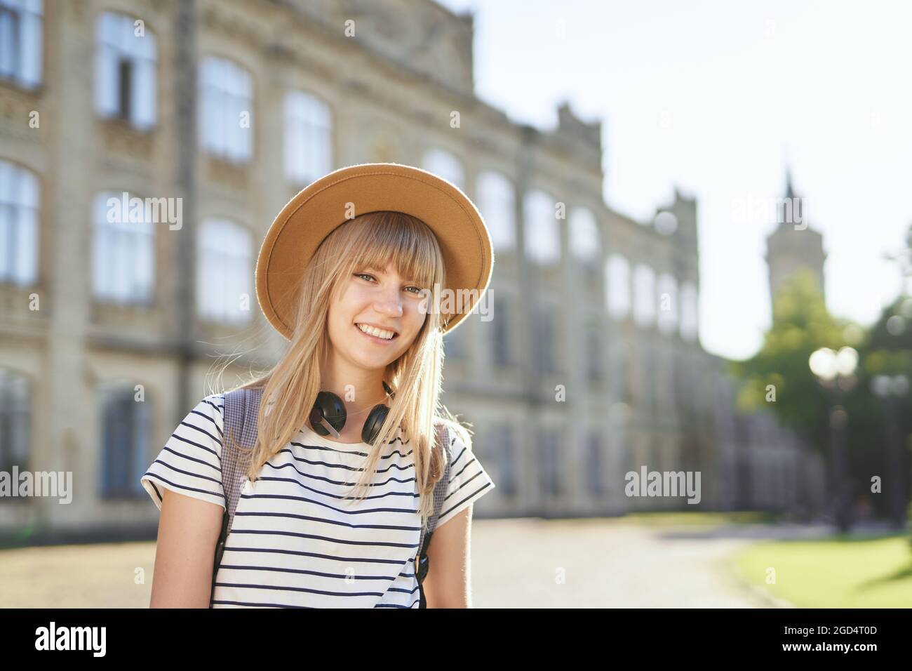 Fröhlich lächelnd junge blonde europäische Universitäts- oder Hochschulstudentin mit braunem Hut auf dem Campus. Nettes Schulmädchen, Studentenportrait oder Bildungskonzept. Hochwertige Bilder Stockfoto
