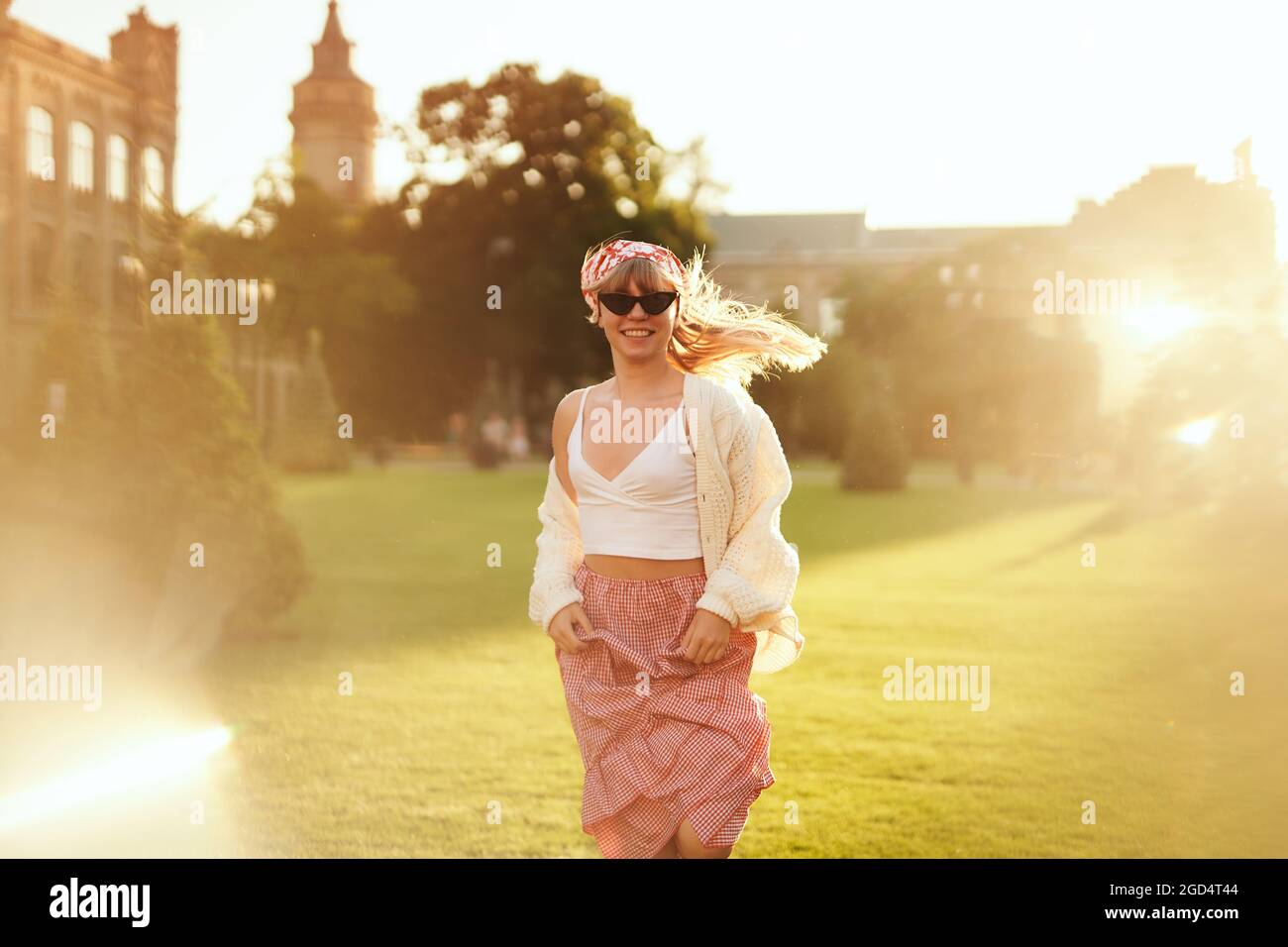 Weibchen läuft in einem grünen Sommerpark zur Kamera. Attraktive blonde Mädchen in roten Rock, weißen Pullover und Kopftuch. Jugendlich im modischen Stil. Urlaub in der Natur, Sommer oder Reisekonzept an sonnigen Tagen Stockfoto