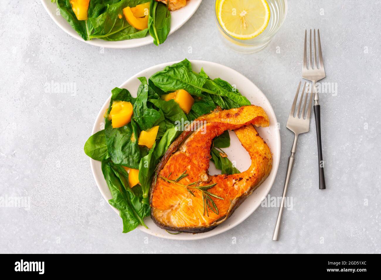 Gebratenes Lachssteak mit Spinat und gelben Tomaten auf einem Teller auf grauem Grund, ein Glas Wasser mit Zitrone, ein herzhaftes Mittagessen mit gesundem rotem Fisch Stockfoto