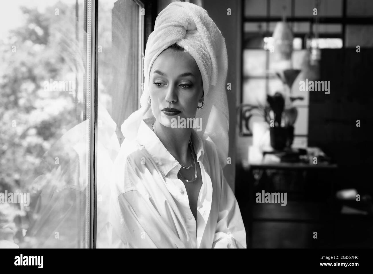 Porträt einer wunderschönen jungen Frau, die weißes Hemd und Turban-Handtuch auf dem Kopf trägt, während sie sich am Fenster entspannt. Stockfoto