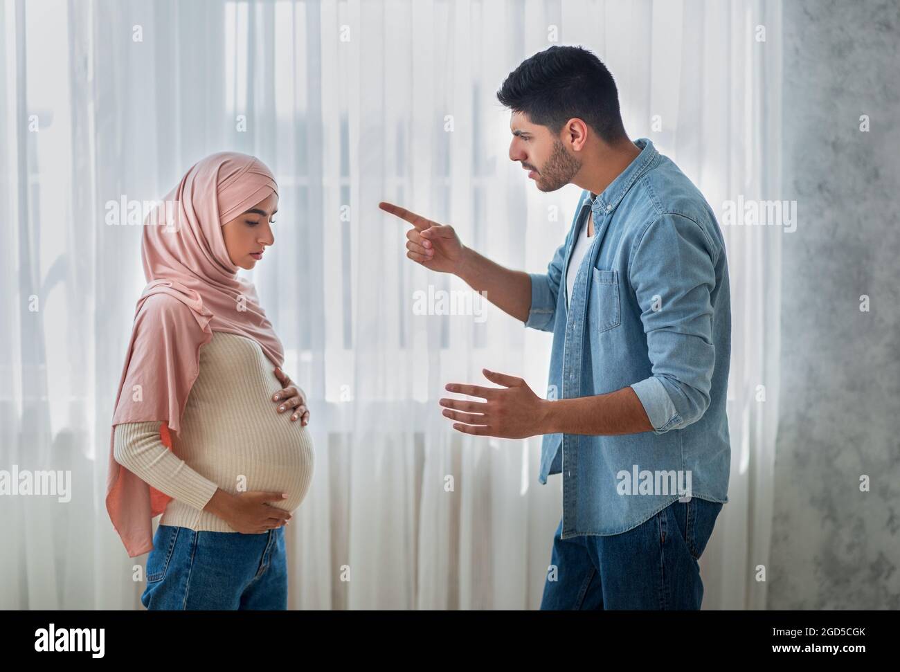 Beziehungsprobleme. Aggressiver arabischer Mann, der mit seiner verärgerten, schwanger-muslimischen Frau zu Hause argumentiert, neben dem Fenster stehend, Seitenansicht. Gestresst erwartungsvoll la Stockfoto