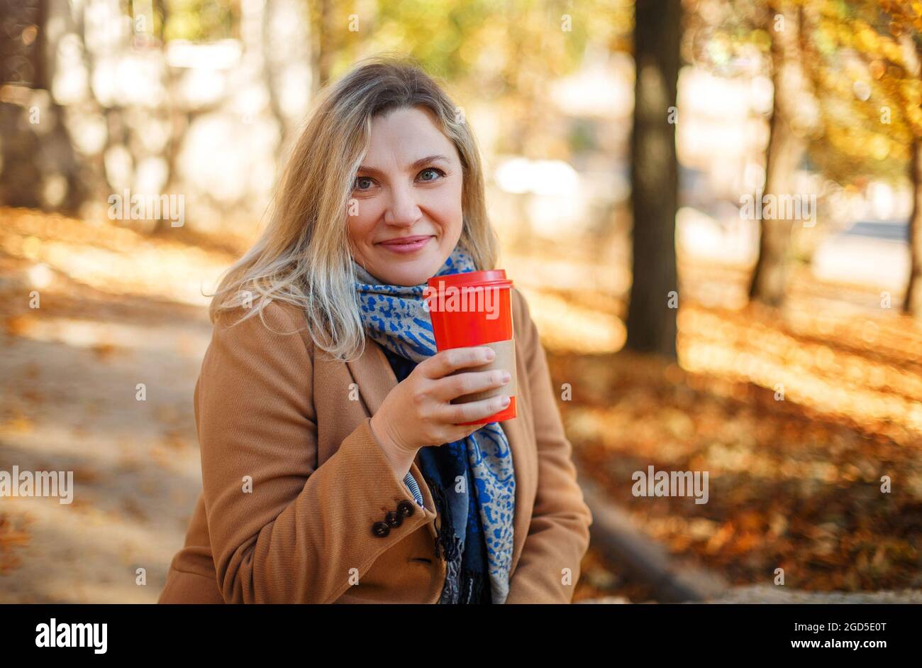 Schöne lächelnde blonde Frau mittleren Alters mit beigefarbenem Mantel und Schal, die am sonnigen Herbsttag im Stadtpark sitzt und ihre Tasse Kaffee zum Mitnehmen genießt. L Stockfoto