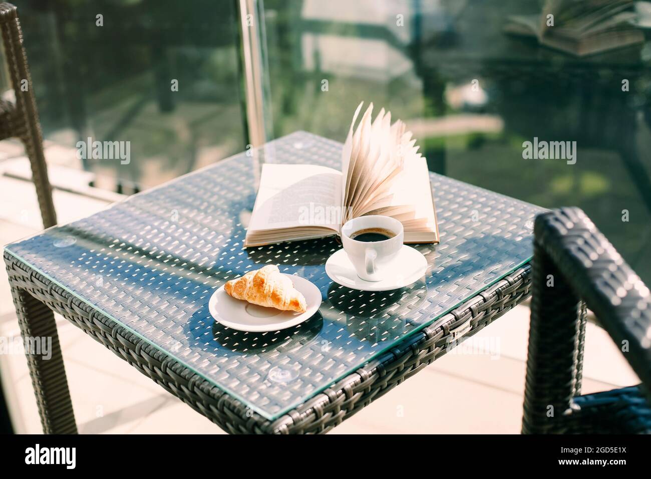 Tasse schwarzen Kaffee, frisch gebackenes Croissant und offenes Buch auf Rattantisch auf dem Hotelbalkon vor verschwommener grüner Resortlandschaft. Morgen bre Stockfoto