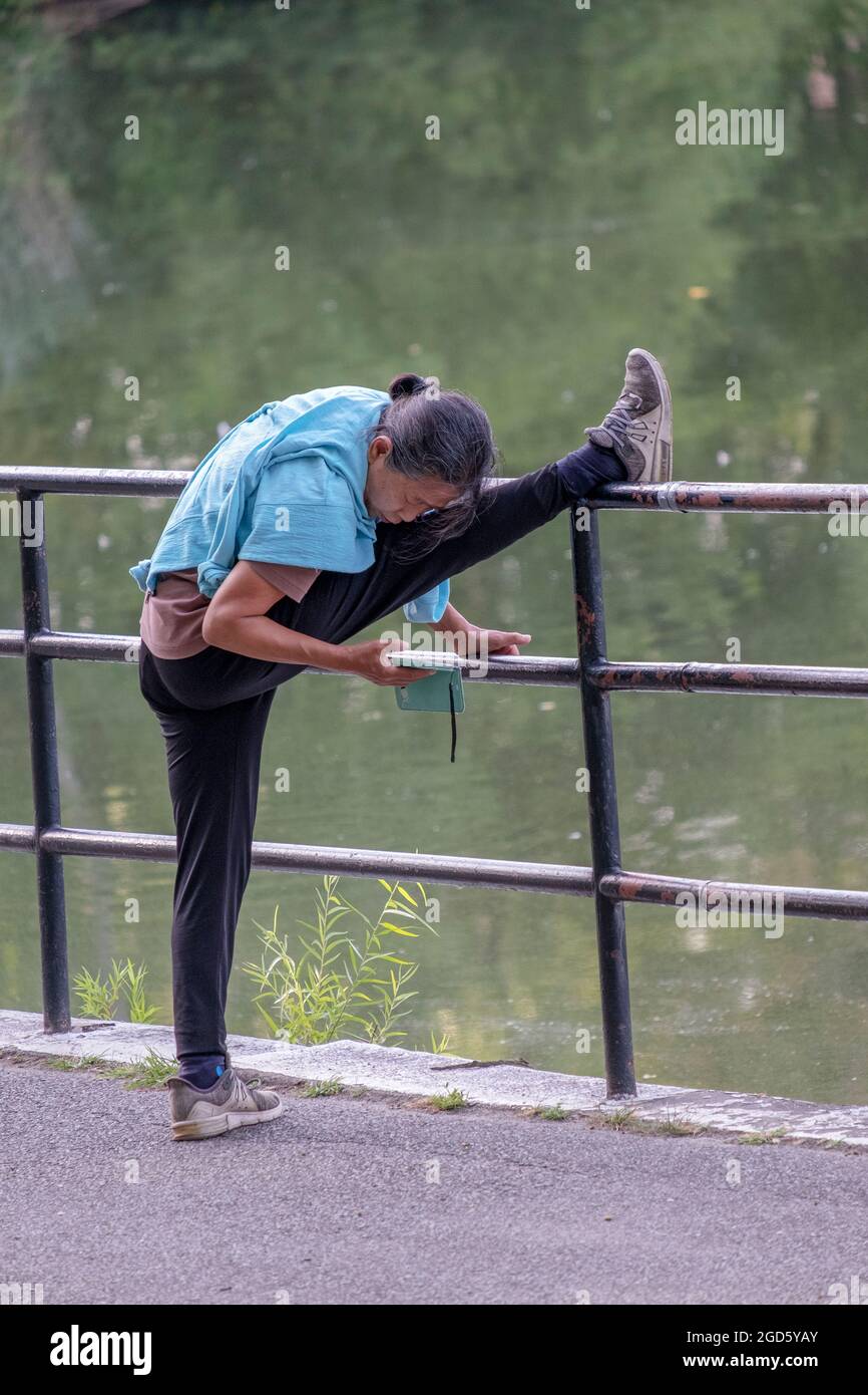 MULTITASKING. Eine Frau streckt ihren Oberschenkel und liest gleichzeitig ihr Handy. Im Kissena Park in Flushing, Queens, New York City. Stockfoto