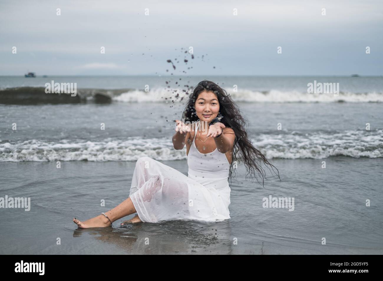Charmante junge mongolische Frau in weißem Kleid sitzt am Strand und spielt mit Sand. Werfen senden Sie auf. Schwarzes langes lockiges Haar. Romantisch. Meerblick Stockfoto
