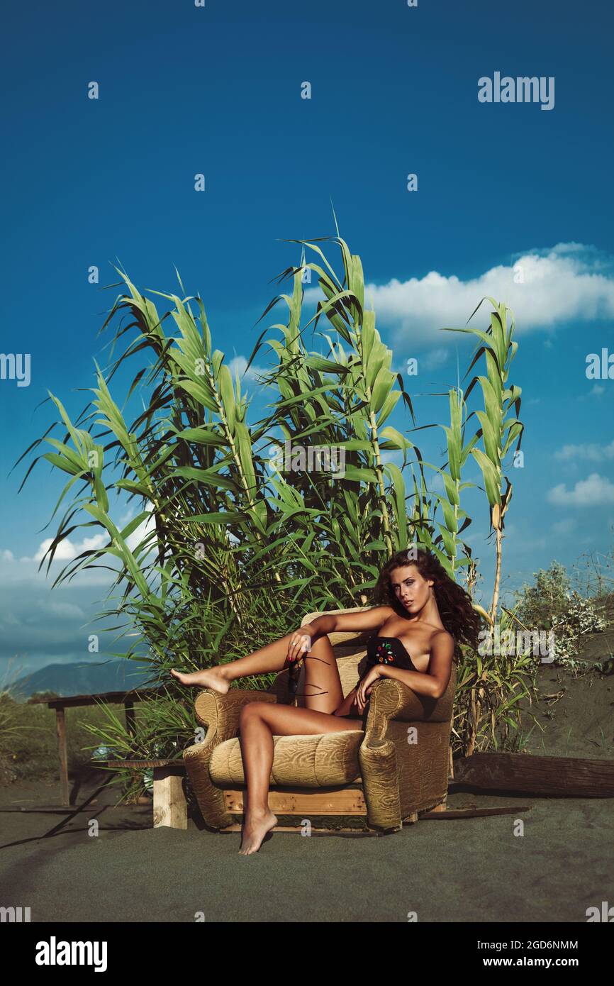 Junge Frau in Bikini und Top sitzen auf einem Sessel am Sandstrand Stockfoto