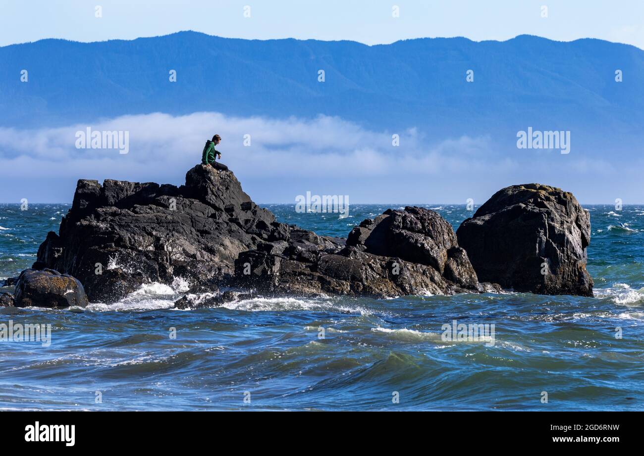 Mann sitzt auf Felsen im French Beach Provincial Park, Vancouver Island, Kanada mit den Bergen des Olympic National Park im Hintergrund. Stockfoto