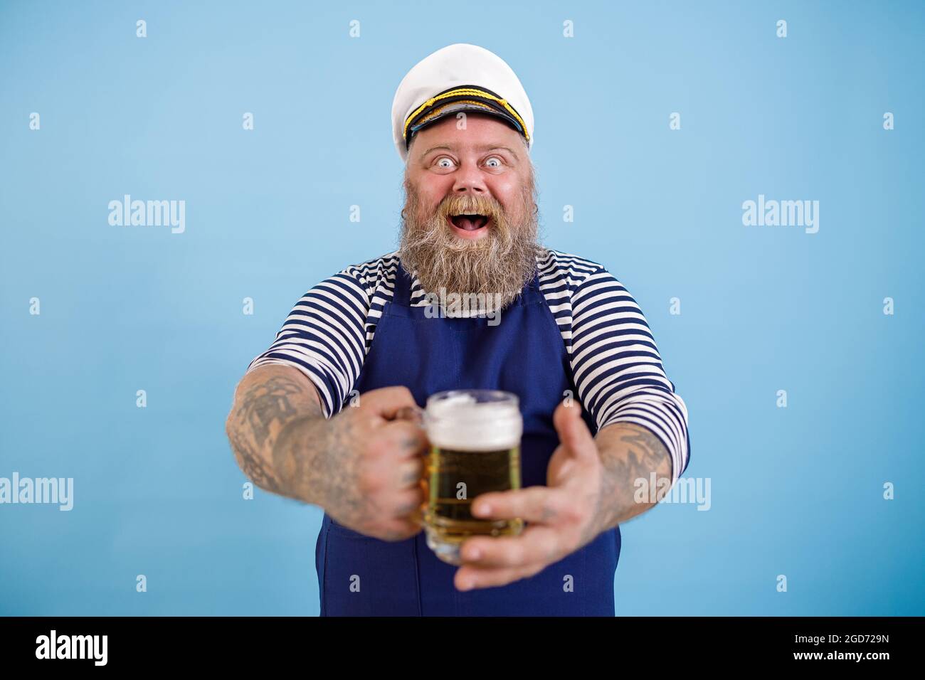 Ein fröhlicher, fetter Mann im Matrosenanzug bietet einen Becher frisches Bier auf hellblauem Hintergrund an Stockfoto