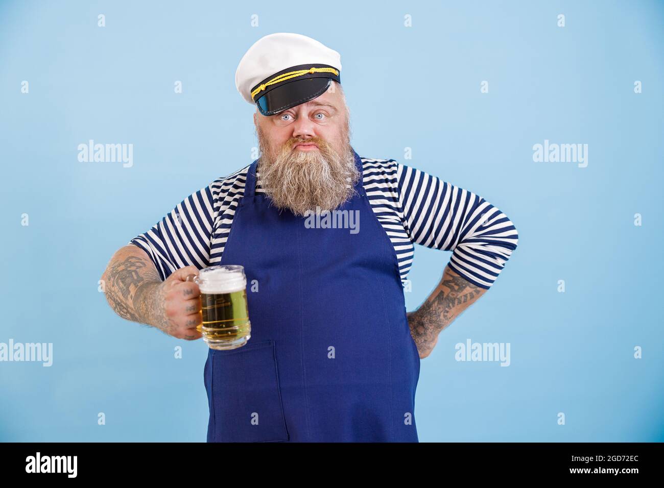 Lustiger Mann in Übergröße im Matrosenanzug hält ein Glas schäumenden Bieres auf hellblauem Hintergrund Stockfoto