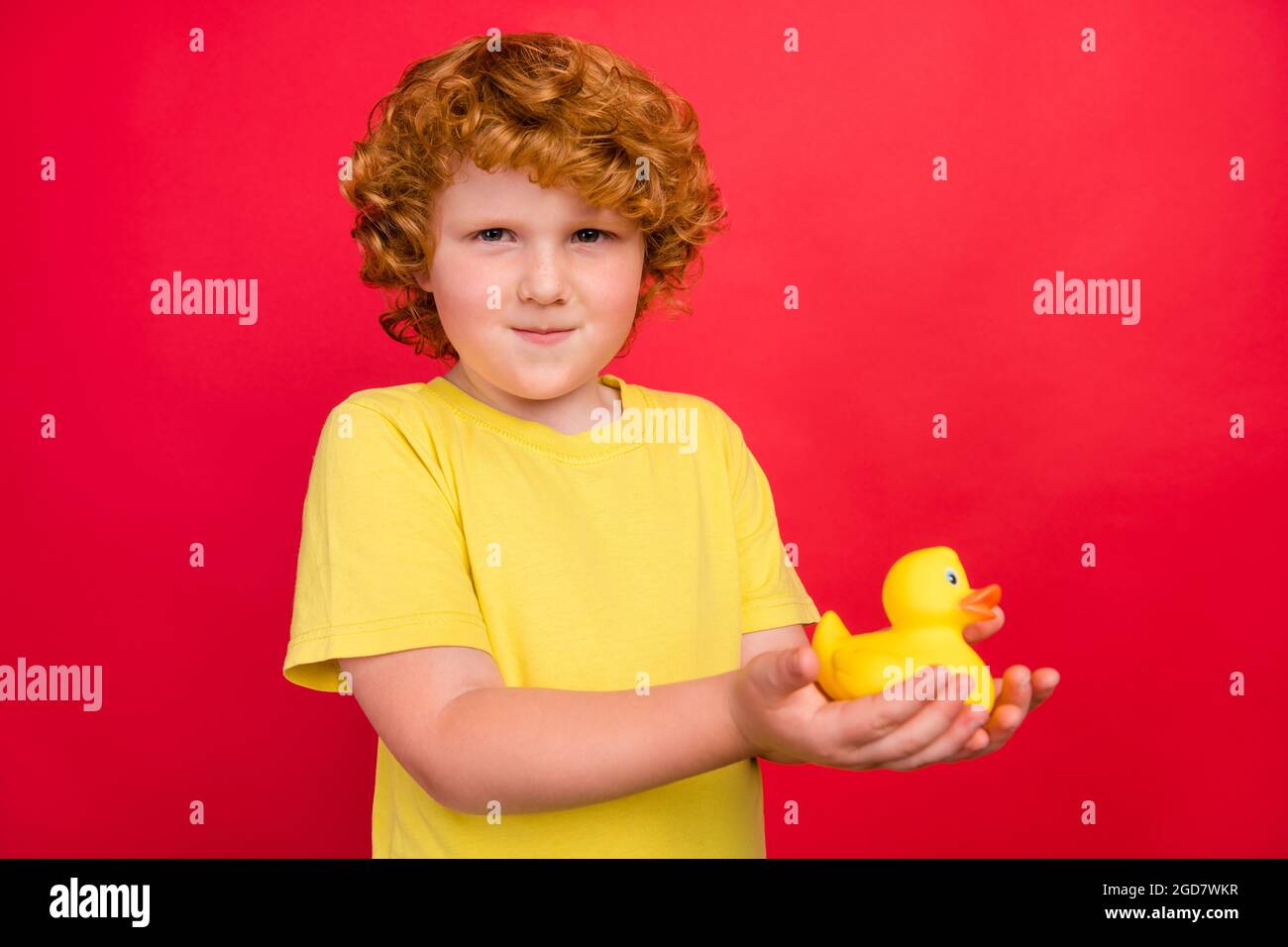 Foto von lustigen ehrlichen Ingwer kleinen Jungen halten Gummi Ente Spielzeug tragen gelbe T-Shirt isoliert roten Hintergrund Stockfoto