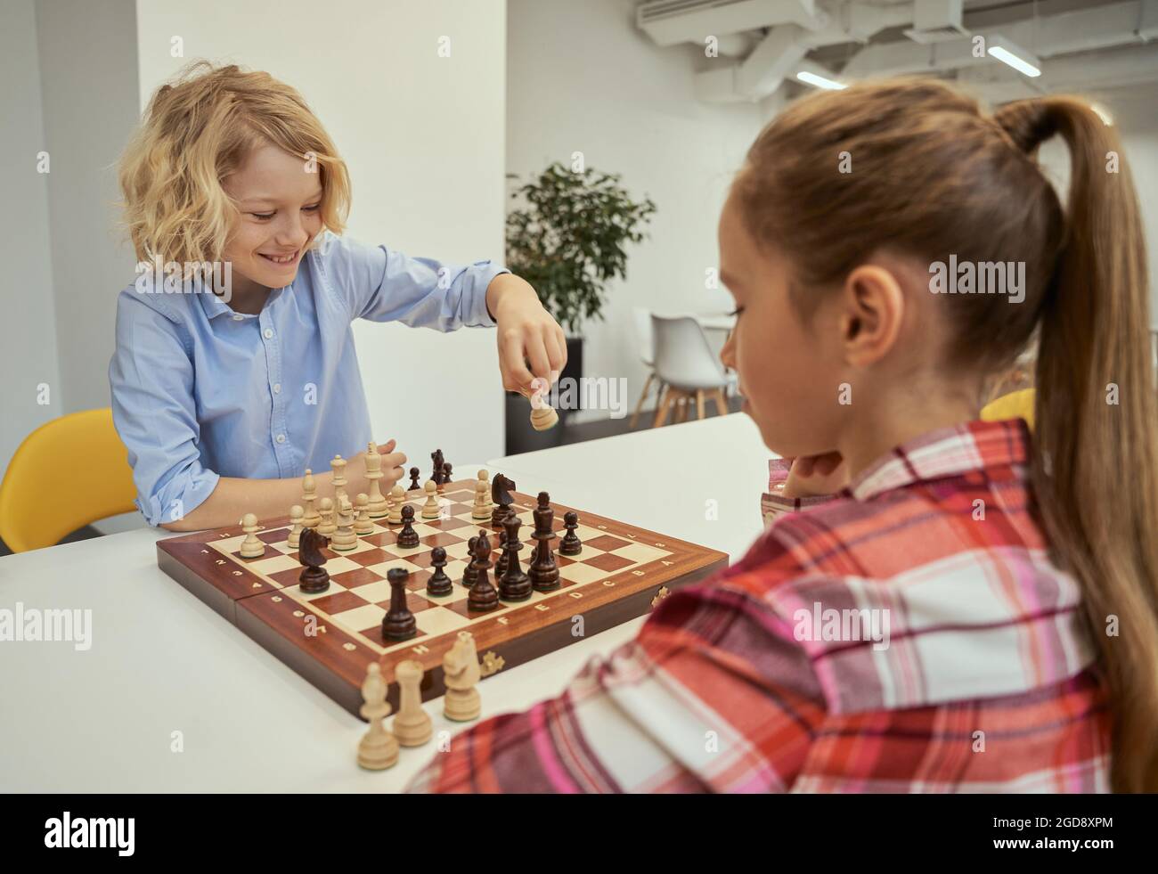 Ein verspielter kleiner kaukasischer Junge lächelt, während er sich bewegt, mit einem Freund Schach spielt und in der Schule zusammen am Tisch sitzt Stockfoto