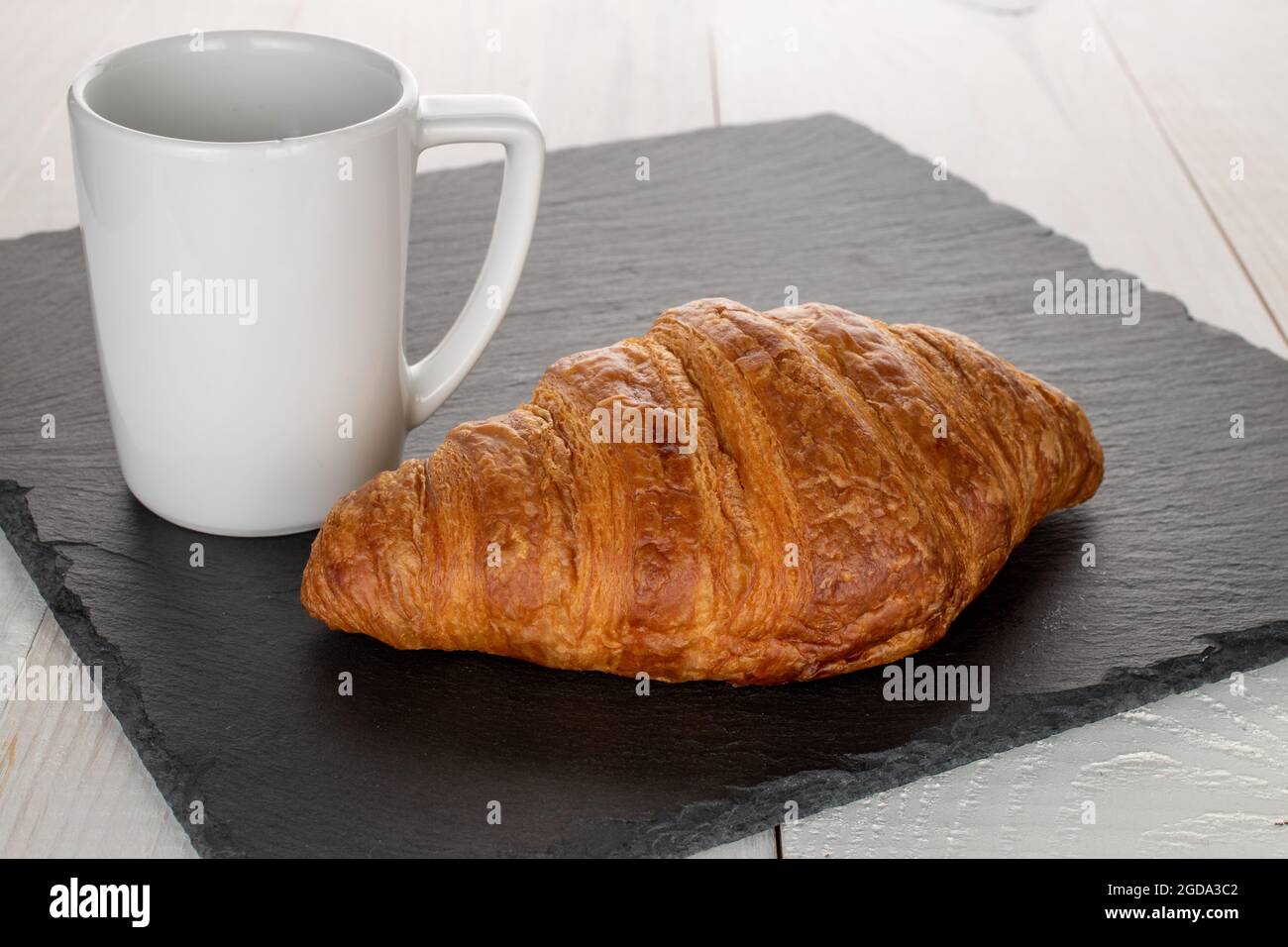 Ein heisses, hausgemachtes Croissant mit einer weißen Tasse, Nahaufnahme, auf einem Schieferbrett. Stockfoto