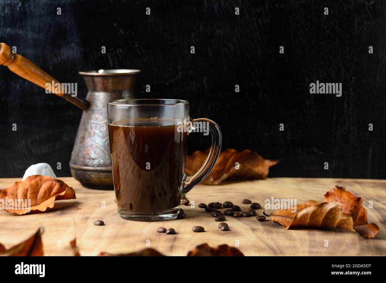Ein Glaskrug aus stark gebrühtem Kaffee auf den Steinen mit Kaffeebohnen und Herbstblättern auf dem Hintergrund einer Kaffeemaschine und dunklem Holz. Selektiv f Stockfoto