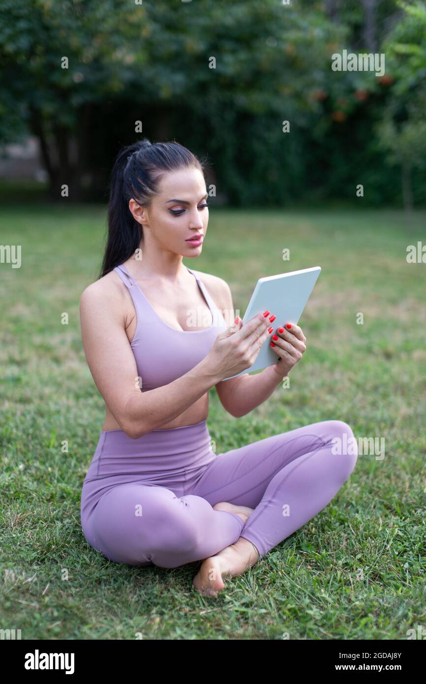 Junge Sportlerin mit digitalem Tablet im Freien, auf Gras sitzend. Mit der intelligenten Fitness-App Stockfoto