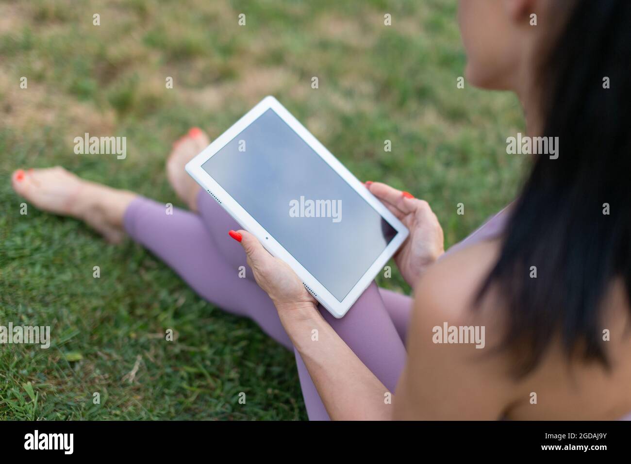 Junge Frau in Sportbekleidung mit intelligenter Fitness-Anwendung auf digitalem Tablet, auf Gras sitzend Stockfoto