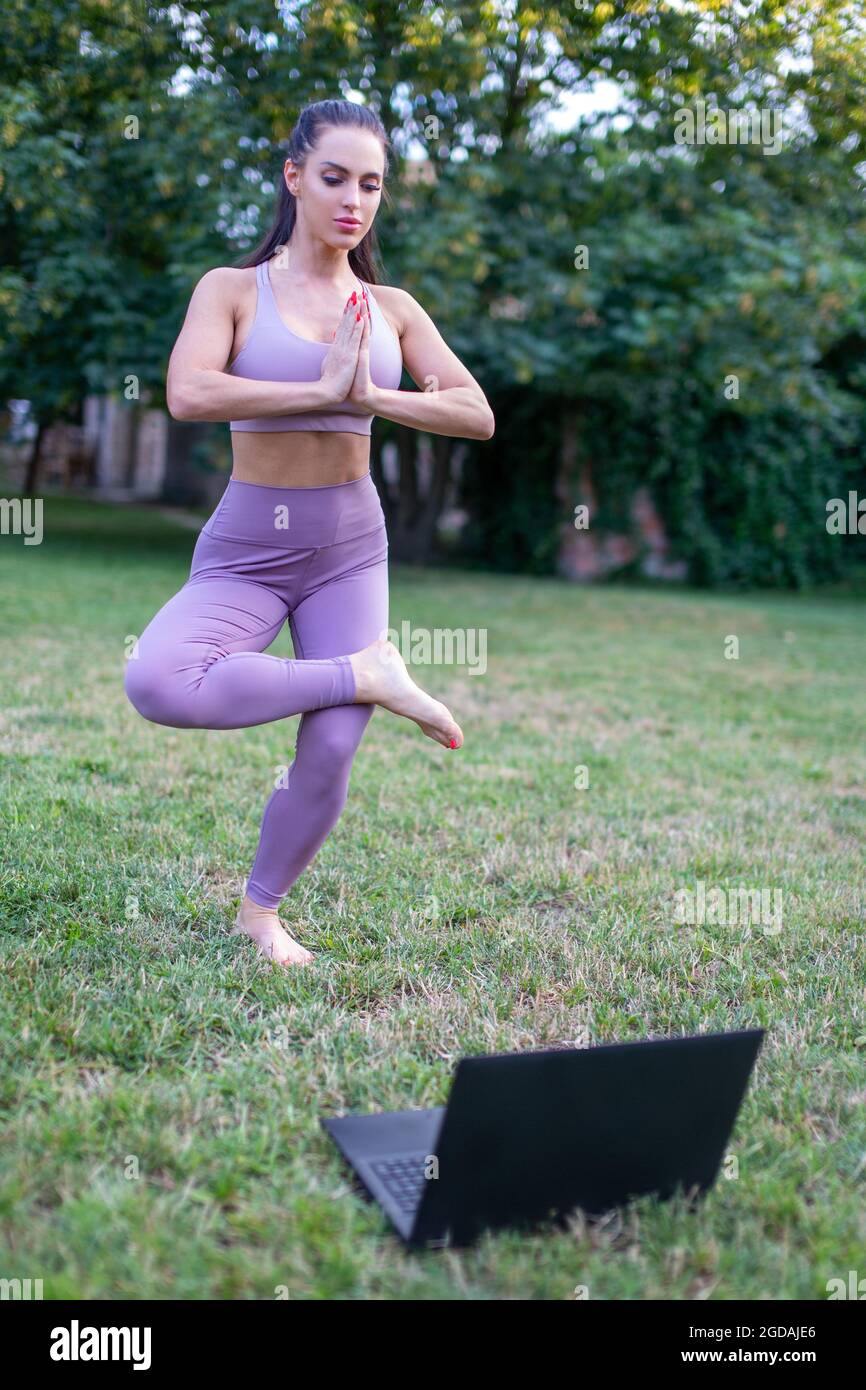 Junge kaukasische Frau, die im Garten stehend Yoga-Pose aus einem Tutorial-Video auf einem Laptop lernt Stockfoto