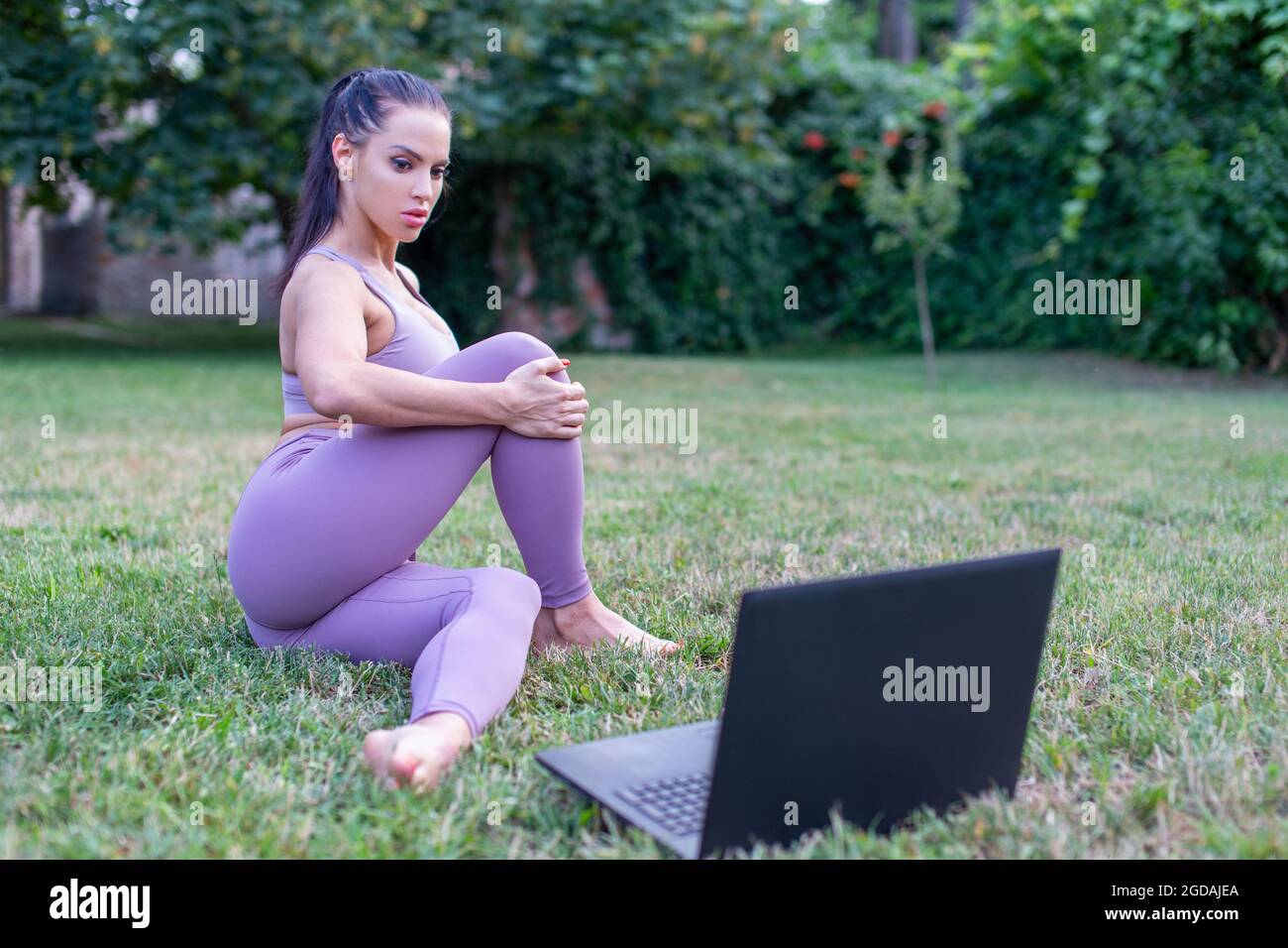 Junge Frau, die sitzende Yoga-Pose aus dem Tutorial-Video auf dem Laptop im Garten lernt Stockfoto