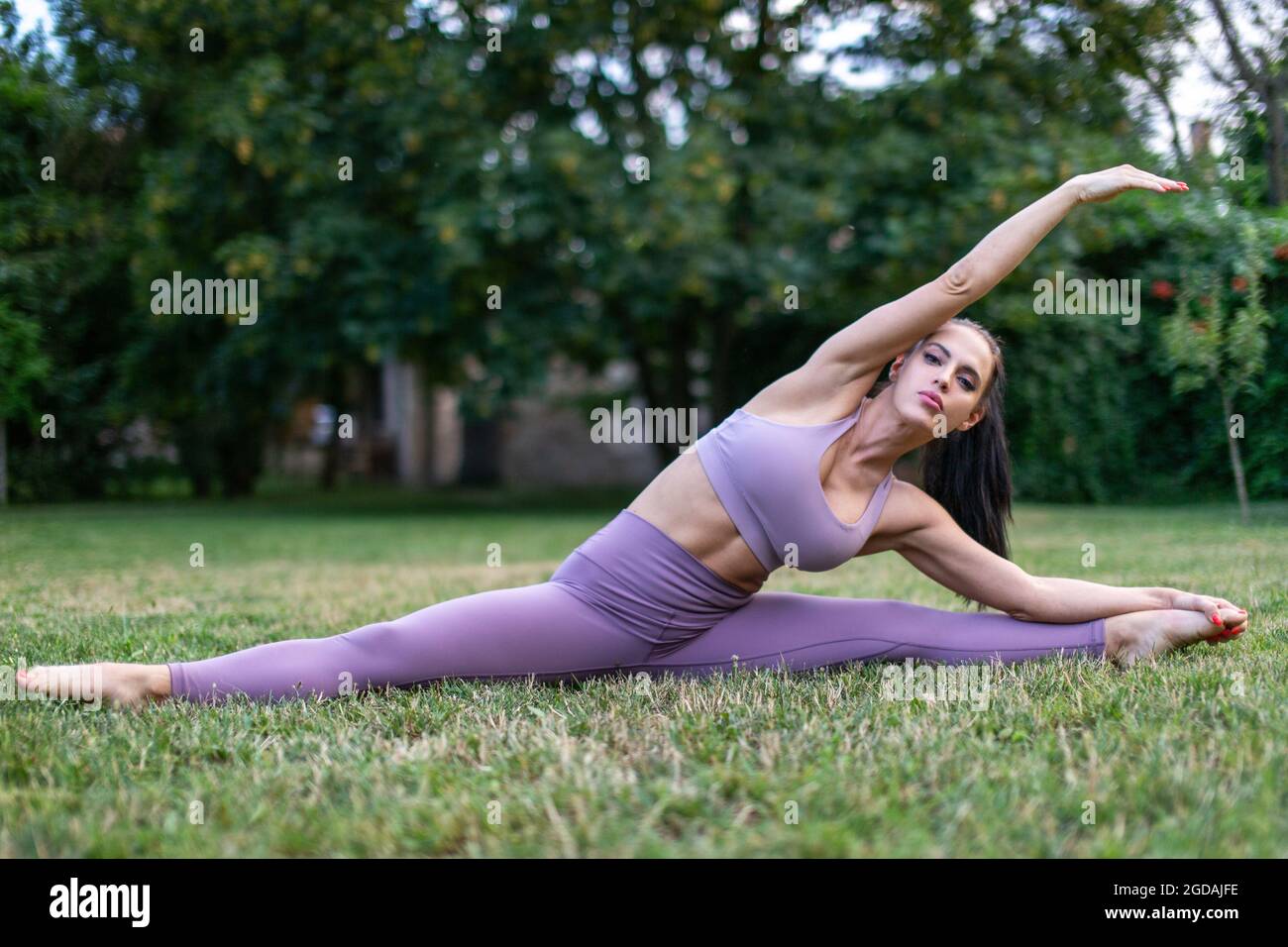 Junge Frau in Sportbekleidung, die sich in gespaltener Haltung im Garten auf Gras, im Freien, ausdehnt Stockfoto