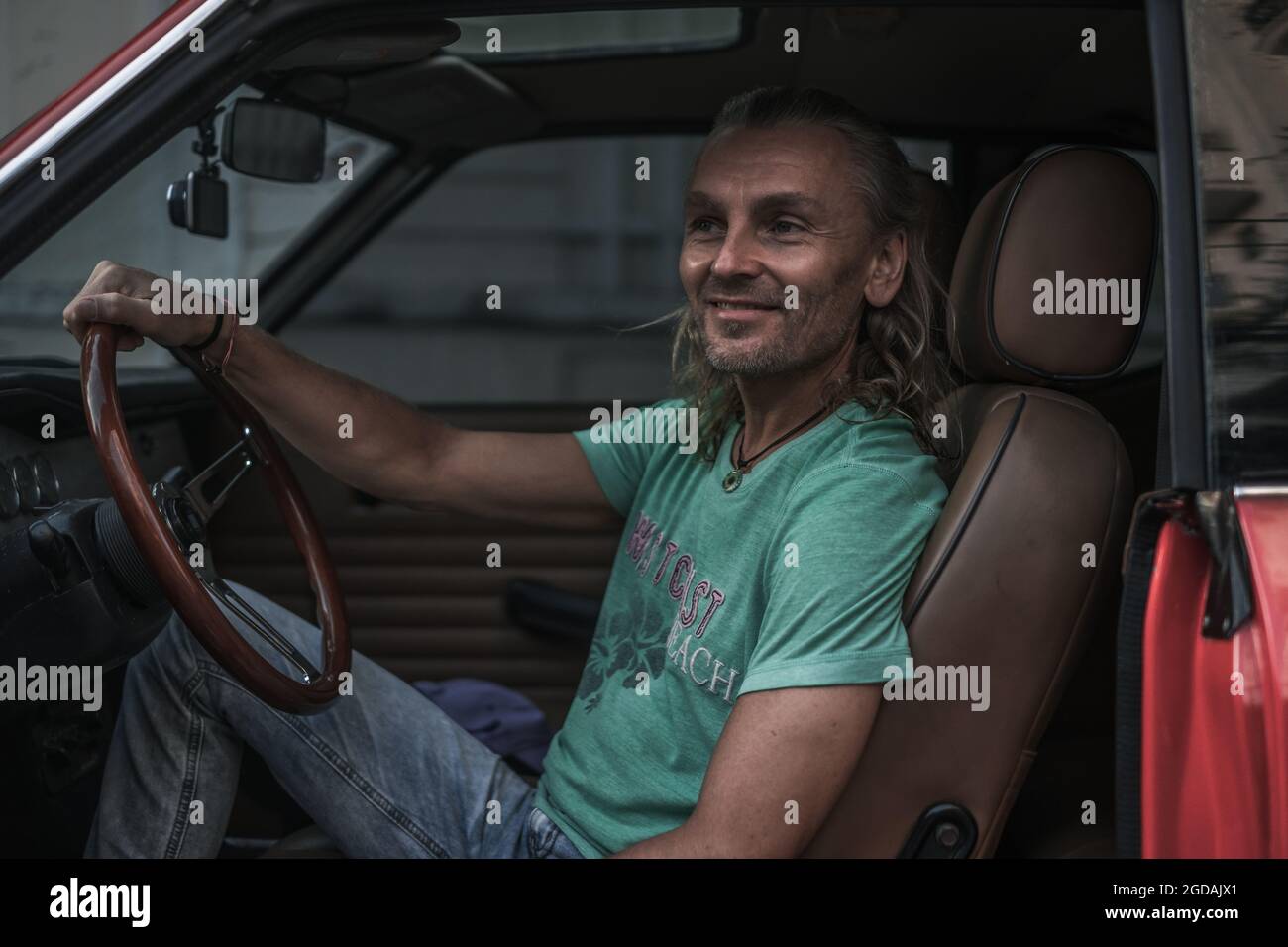 Porträt eines reifen hübschen Mannes, der in einem roten Auto im alten Stil sitzt, das Lenkrad hält und lächelt. Lange graue Haare. Rotes Auto im Stil der 1970er Jahre. Hohe Qualität Stockfoto