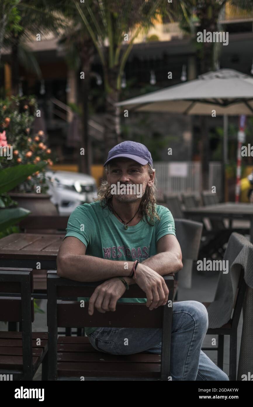 Porträt eines alten Mannes, der auf der Sommerverande des Restaurants sitzt. Lange graue Haare. Street-Style. Hippie. Hochwertige Fotos Stockfoto