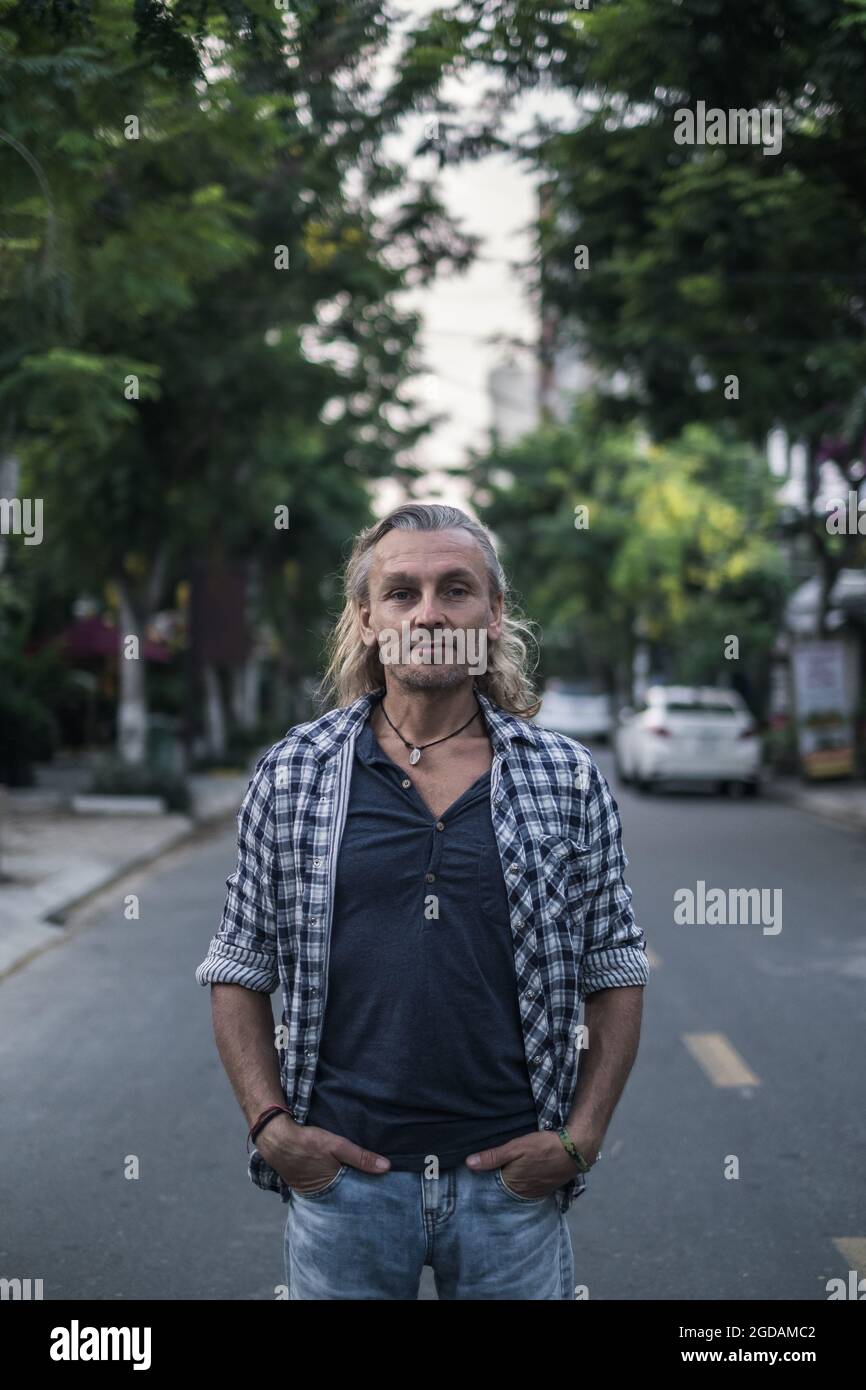 Porträt eines älteren, gutaussehenden Mannes in Hemd und Jeans, der auf der Straße in der Stadt steht. Lange graue Haare. Hochformat. Hochwertige Fotos Stockfoto