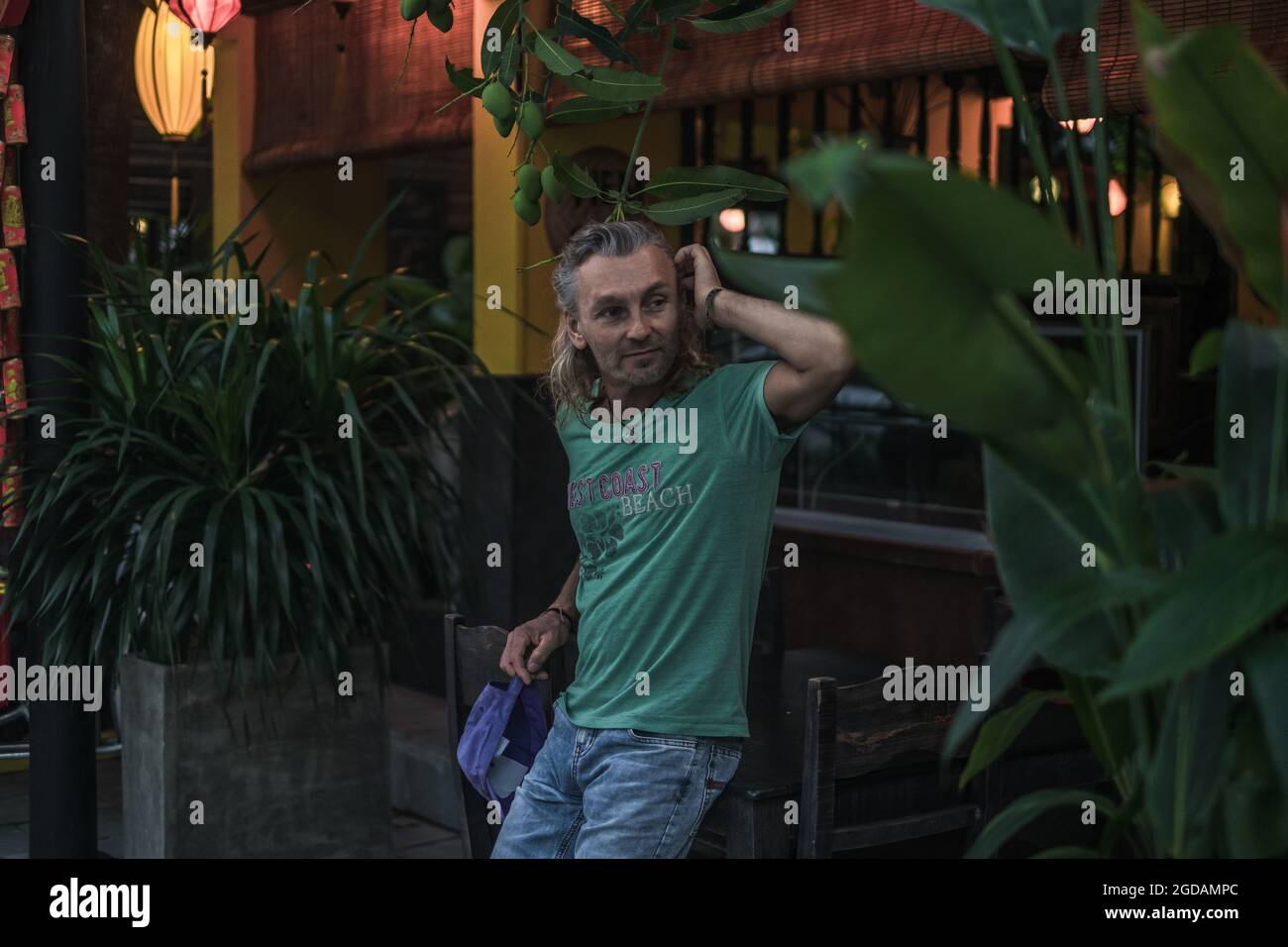 Porträt eines reifen Mannes in T-Shirt, Jeans stehend und mit dem Kopf. Lange graue Haare. Chinesische Laternen. Abend. Tropische Bäume im Hintergrund. Stockfoto