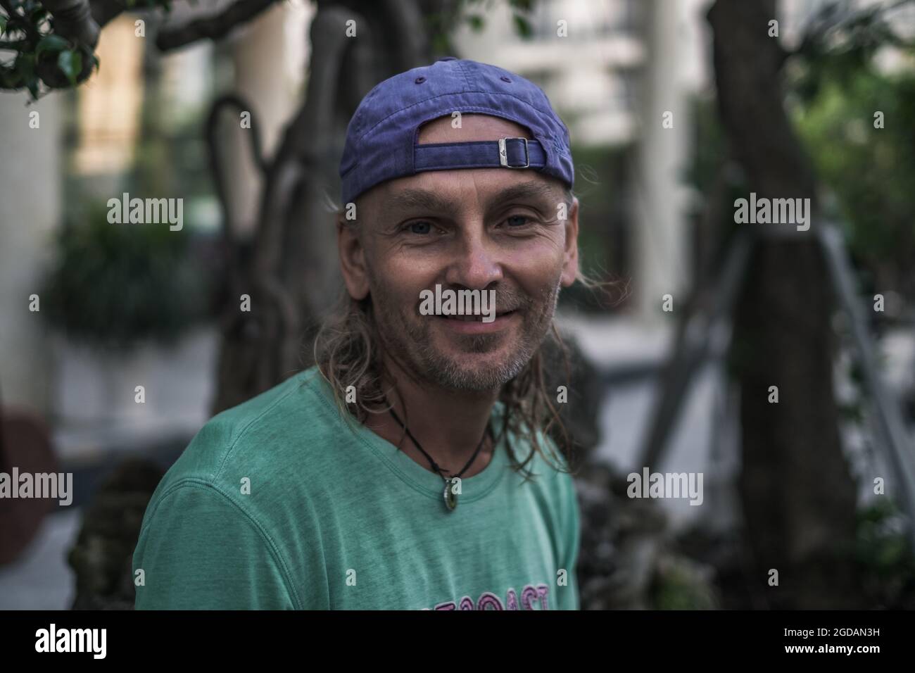 Nahaufnahme eines lächelnden älteren Mannes mit langen grauen Haaren und grünem T-Shirt und Mütze. Obdachlose Person. Hippie-Style. Hochwertige Fotos Stockfoto