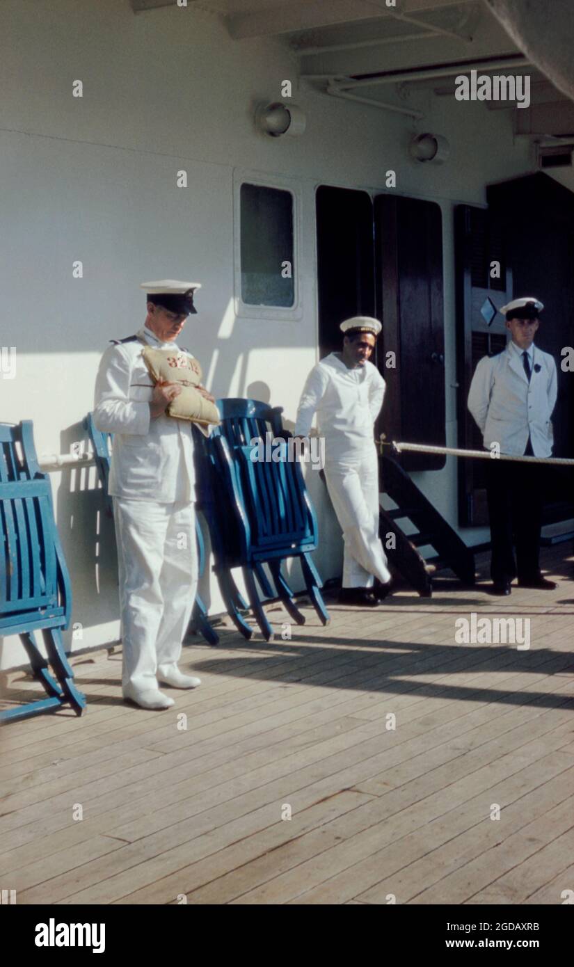 Ein uniformierte Offizier, der darlegt, wie man eine Rettungsweste oder ein persönliches Flotationsgerät auf dem Kreuzfahrtliner SS Maasdam der Holland America Line anlegt und trägt, während er 1959 in New York, USA, im Dock war. Hier würde ein Leinwandbeutel flexible wasserdichte Zellen enthalten, die mit Kapok gefüllt sind. Das Gerät ist nutzbringend nummeriert (hier 32.12), um eine Organisation an Bord des Schiffes und jeden Rettungsversuch zu unterstützen. Dieses Gerät (PFD, Rettungsweste, Rettungsring, Rettungsgurt, Mae West, Rettungsweste, Rettungsschwimmer, Korkjacke, Schwimmhilfe oder Schwimmanzug) wird getragen, um Ertrinken zu verhindern – ein Vintage-Foto aus den 1950er Jahren. Stockfoto Ein uniformierte Offizier, der darlegt, wie man eine Rettungsweste oder ein persönliches Flotationsgerät auf dem Kreuzfahrtliner SS Maasdam der Holland America Line anlegt und trägt, während er 1959 in New York, USA, im Dock war. Hier würde ein Leinwandbeutel flexible wasserdichte Zellen enthalten, die mit Kapok gefüllt sind. Das Gerät ist nutzbringend nummeriert (hier 32.12), um eine Organisation an Bord des Schiffes und jeden Rettungsversuch zu unterstützen. Dieses Gerät (PFD, Rettungsweste, Rettungsring, Rettungsgurt, Mae West, Rettungsweste, Rettungsschwimmer, Korkjacke, Schwimmhilfe oder Schwimmanzug) wird getragen, um Ertrinken zu verhindern – ein Vintage-Foto aus den 1950er Jahren. Stockfoto