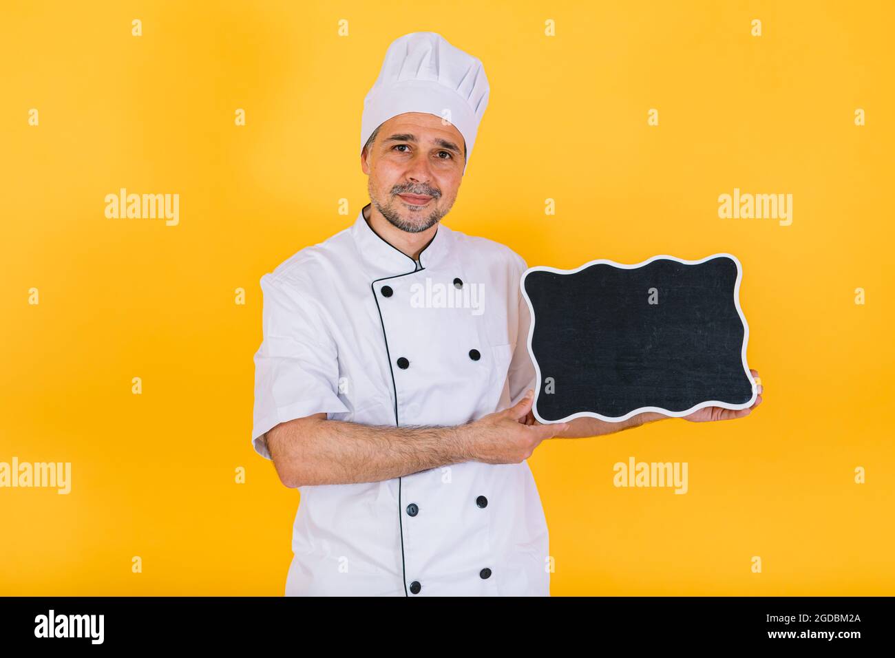 Chefkoch trägt weiße Küchenmütze und Jacke mit schwarzer Kreidetafel auf gelbem Hintergrund Stockfoto