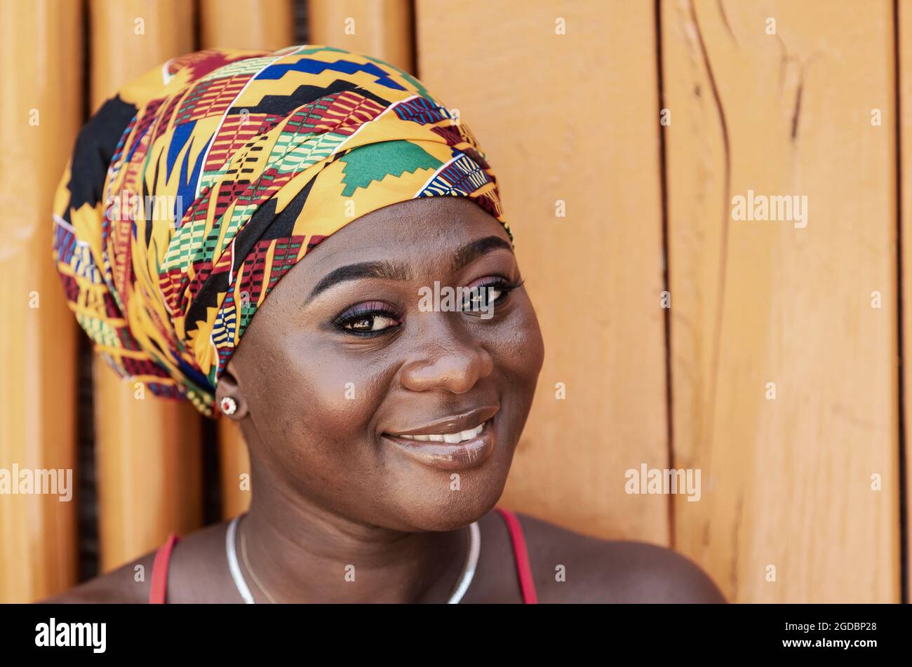 Afrikanische Frau mit einem hoffnungsvollen glücklichen Lächeln und traditionellem Kopfschmuck, die gegen eine bemalte Holzwand im tropischen Dorf Keta Ghana, West, steht Stockfoto