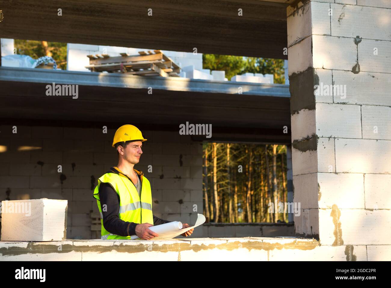 Ein Bauingenieur und Konstrukteur in einem gelben Hardhat studiert eine Zeichnung eines Gebäudes auf einer Baustelle. Ein Haus aus porösem Betonblock Stockfoto