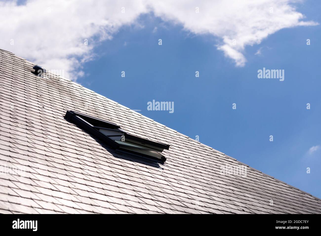 Ein Porträt eines geöffneten Dachfensters zur Belüftung in einem Schieferdach an einem sonnigen Tag mit einem blauen Himmel mit weißen Wolken. Sie können auch eine Ventilation sehen Stockfoto