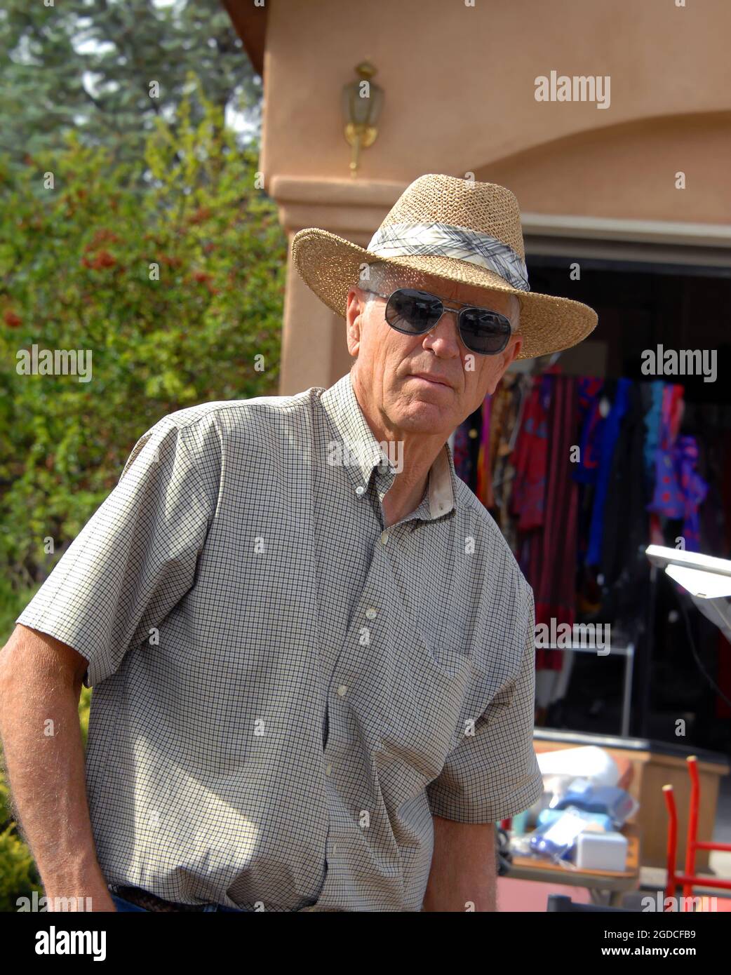 Reifer Mann hält in seinem Shopping in die Kamera zu schauen. Er ist in der garage Saling und trägt ein Stroh Hut, Sonnenbrille und Plaid Shirt. Stockfoto