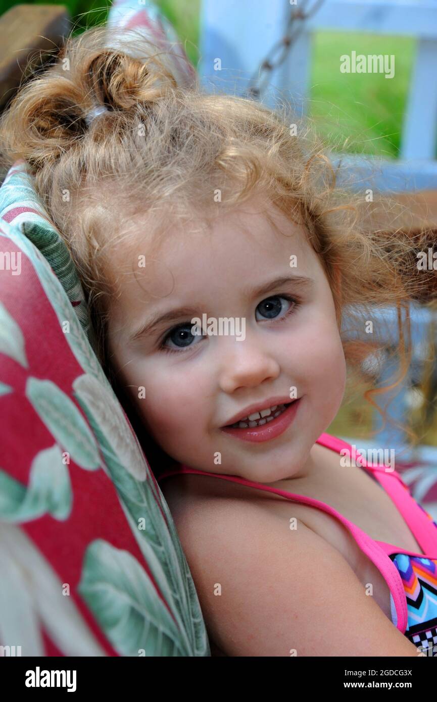 Das kleine Mädchen genießt es, auf der Veranda zu schwingen. Sie schaut seitwärts mit dem Kopf auf dem Schaukelkissen. Locken sind ihrem Pferdeschwanz entkommen. Stockfoto