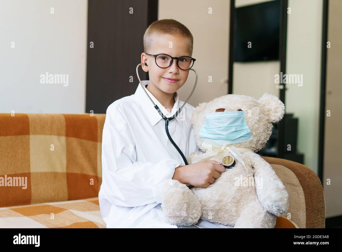 Blick auf ein Kind, das zu Hause im Sonnenlicht einen Arzt oder eine Krankenschwester mit einem Teddybären spielt. Happy Boy hört einem Spielzeug Stethoskop zu. Rollenspiel eines verspielten Kerls. In einer Brille und einem Gewand mit Schutzmaske Stockfoto