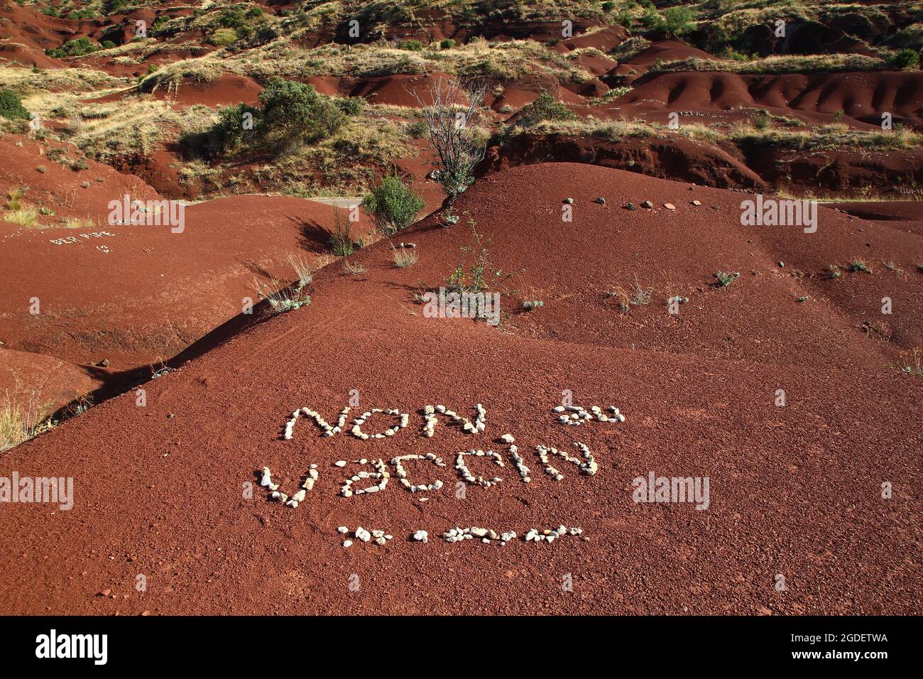 Auf dem roten Boden der Ufer des Salagou-Sees haben Aktivisten Kieselsteine verwendet, um eine feindliche Botschaft bezüglich der Impfung gegen Covid 19 (Frankreich) zu schreiben. Stockfoto