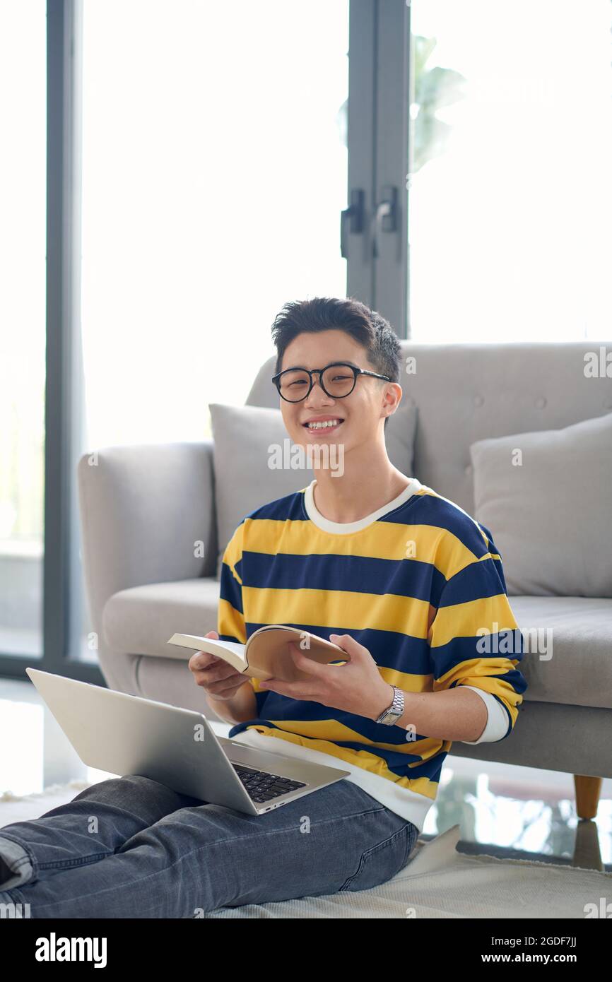 Freiberuflicher Arbeitstag. Lächelnder, tausendjähriger Student, der von zu Hause aus auf dem Teppich im Wohnzimmer sitzt und studiert. Stockfoto