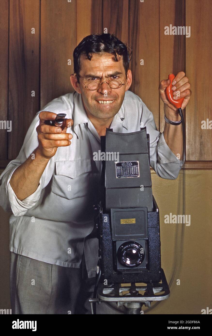 Studiofotografie in Amerika in den 1950er Jahren – ein Fotograf konzentriert sich hart und drückt mit seiner riesigen Graflex-Kamera einen Auslöserauslöser für die Glühbirne. Die Lampe (ein abnehmbares pneumatisches Gummigerät an einem Kabel, das mit der Kamera verbunden ist) bedeutete, dass der Bediener Verwacklungen der Kamera vermeiden konnte. Außerdem war es sehr nützlich, die Lampe gedrückt zu halten und das Objektiv offen zu halten, um Langzeitbelichtungen zu ermöglichen. Graflex war ein amerikanischer Kamerahersteller. Dieses Bild stammt aus einer alten amerikanischen Kodak Amateur-Farbtransparenz – einem Vintage-Foto aus den 1950er Jahren. Stockfoto