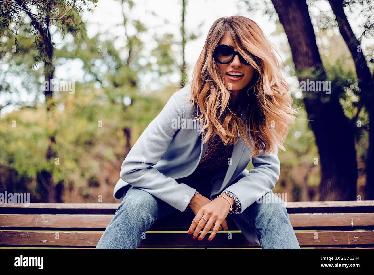 Stilvolle Frau mit langen blonden Haaren tragen Sonnenbrillen sitzen auf einer Parkbank, Porträt Stockfoto