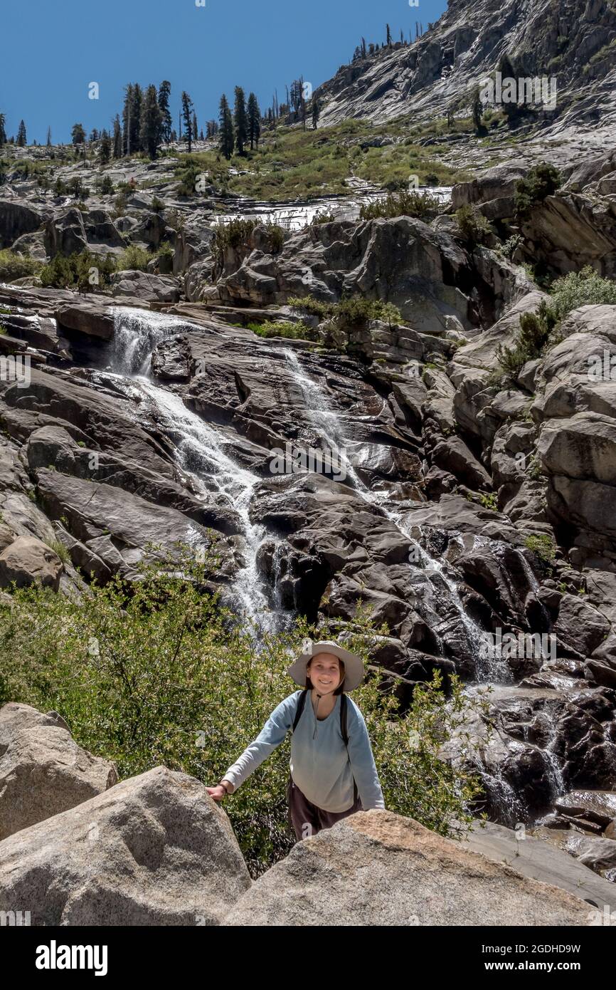 Ein Teenager-Mädchen lächelt beim Wandern im Sequoia National Park für ein Foto mit dem unteren Teil des Tokopah Falls Wasserfalls. Stockfoto