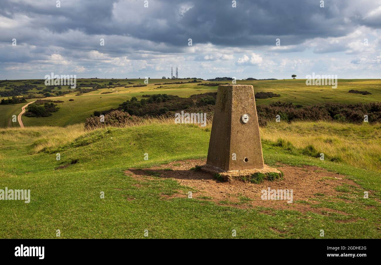 Der Triangulationspunkt auf Cleeve Hill mit Cleeve Common, den drei Telecom Masten und der Gedenkmauer im Hintergrund, Gloucestershire, England Stockfoto