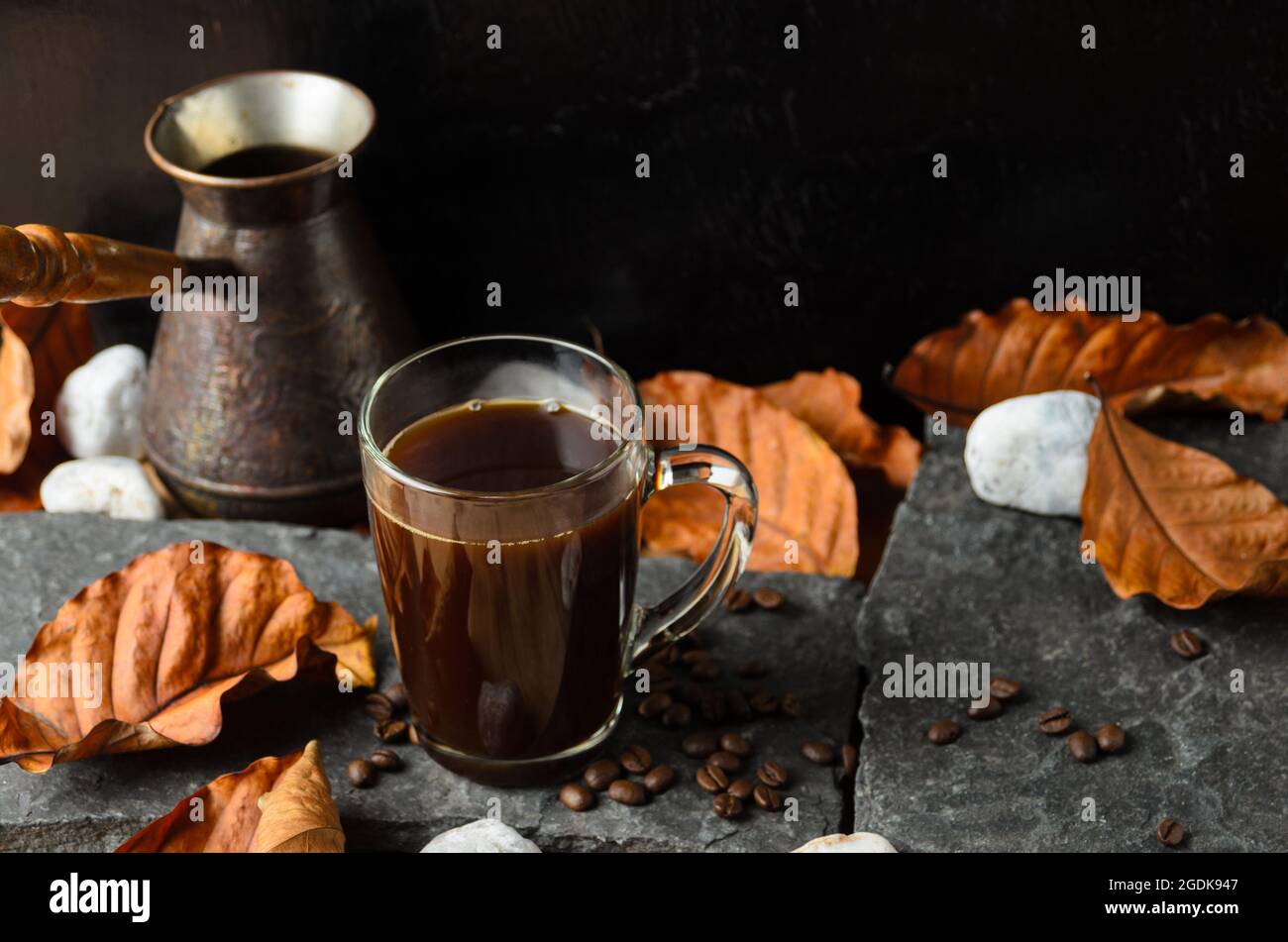 Ein Glaskrug aus stark gebrühtem Kaffee auf den Steinen mit Kaffeebohnen und Herbstblättern auf dem Hintergrund einer Kaffeemaschine und dunklem Holz. Selektiv f Stockfoto