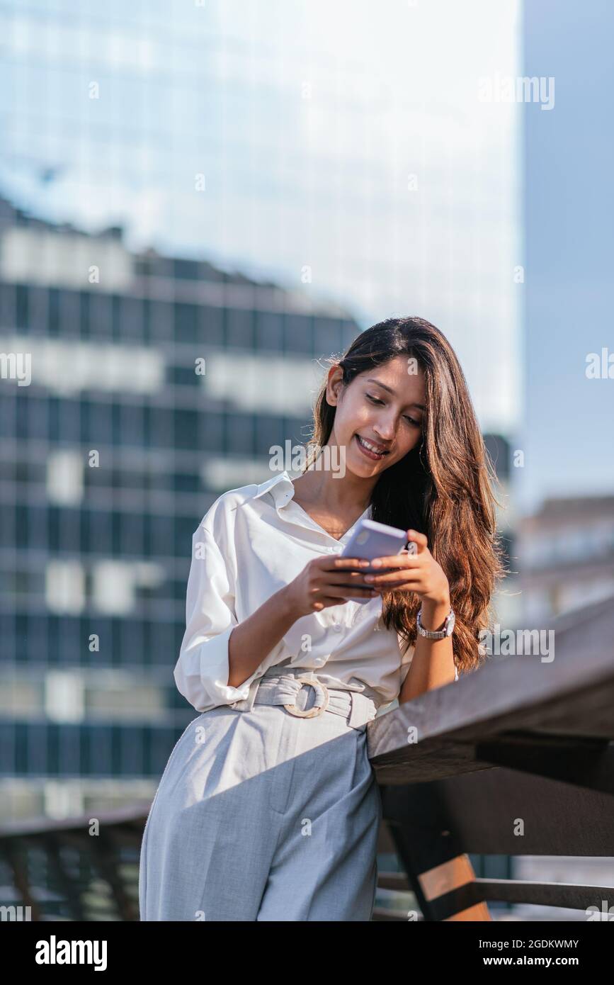 Vertikales Porträt einer attraktiven indischen Frau, die mit ihrem Smartphone eine Sprachnachricht sendet. Sie lehnt sich an das Geländer einer Brücke Stockfoto