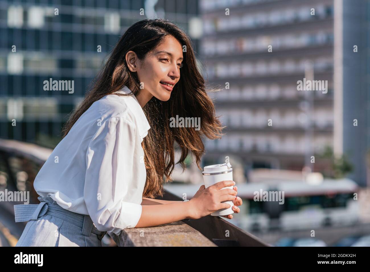 Horizontales Porträt einer attraktiven jungen indischen Geschäftsfrau, die auf einem Balkongeländer liegt. Sie hält einen wiederverwendbaren Kaffeebecher und hat Bürogebäude im Hintergrund Stockfoto
