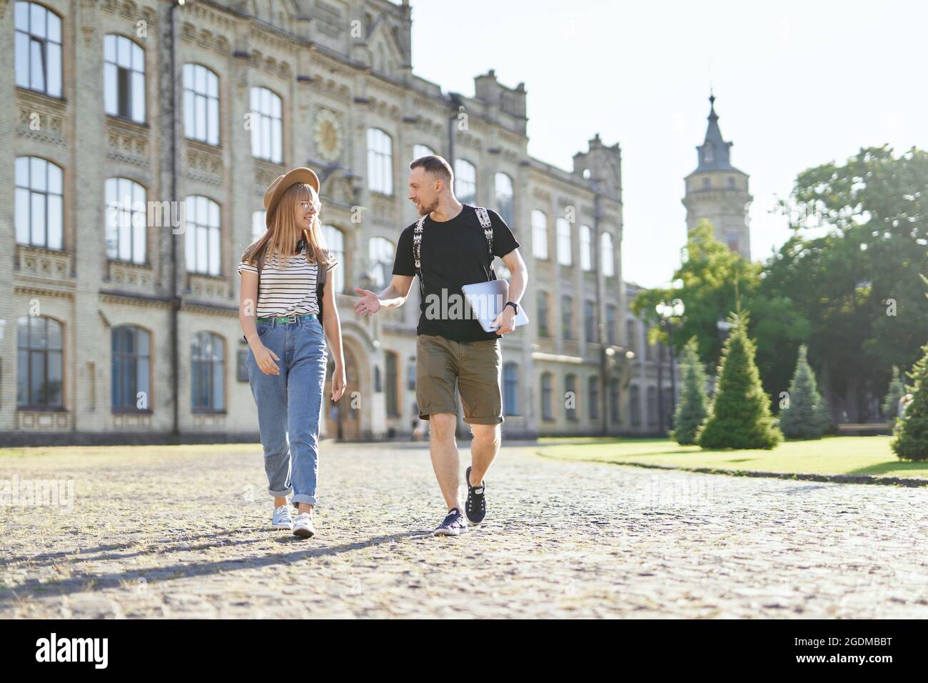 Zwei fröhliche Studenten, männlich und weiblich, mit Rucksäcken, die am Universitätscampus den Daumen nach oben heben und einen Laptop halten. Liebenswert erfolgreiche Studenten paar lächelnd zeigt Daumen nach oben Zeichen an der Kamera Stockfoto